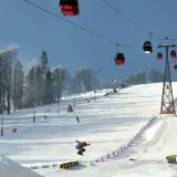Widok na stok narciarski z kolejką linową, snowboardzistą w powietrzu i drzewami w tle.
