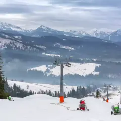 Ośnieżony stok narciarski z armatkami śnieżnymi i wyciągiem krzesełkowym w tle, na tle gór.