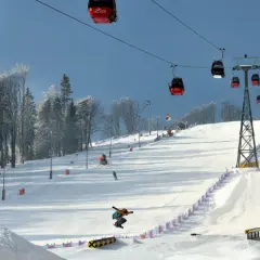 Widok na stok narciarski z kolejką linową, snowboardzistą w powietrzu i drzewami w tle.