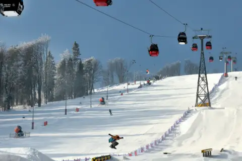 Widok na stok narciarski z kolejką linową, snowboardzistą w powietrzu i drzewami w tle.