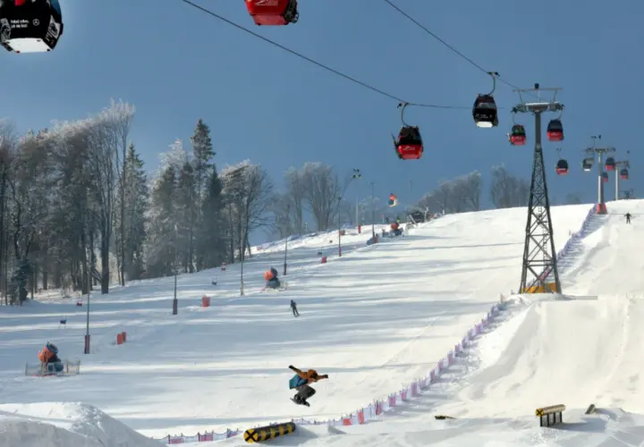 Widok na stok narciarski z kolejką linową, snowboardzistą w powietrzu i drzewami w tle.