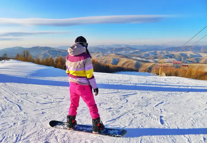 Snowboardzistka w różowych spodniach i kolorowej kurtce na tle górskiego krajobrazu.