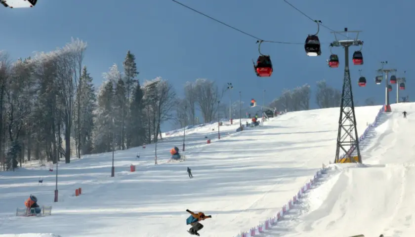 Widok na stok narciarski z kolejką linową, snowboardzistą w powietrzu i drzewami w tle.