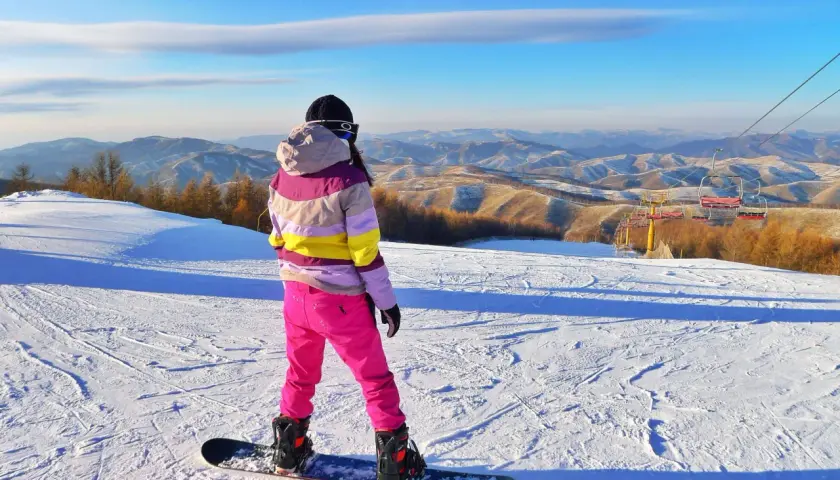 Snowboardzistka w różowych spodniach i kolorowej kurtce na tle górskiego krajobrazu.